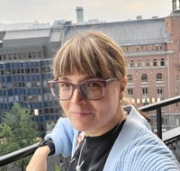 Katja Loikkanen posing for a picture on a balcony while wearing eyeglasses and a black t-shirt with a light-blue cardigan.
